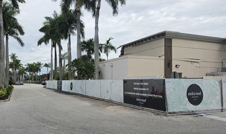 Construction site on a street with palm trees, fenced perimeter, and a beige building behind.