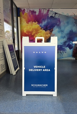 A white A-frame sign with blue panel reading Volvo Vehicle Delivery Area Schumacher indoors against colorful mural.